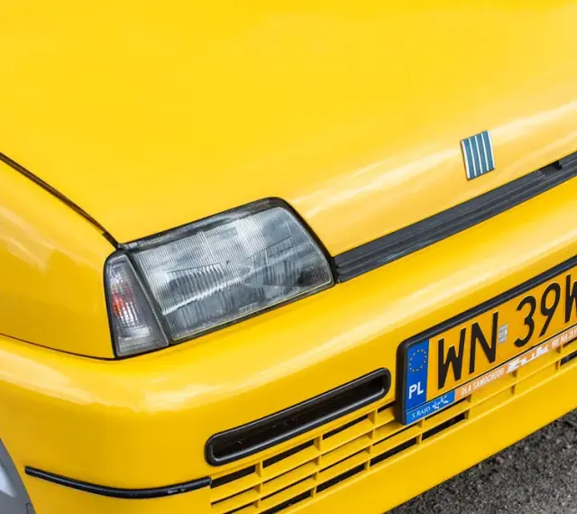 Żółte tablice rejestracyjne na błyszczącym, żółtym samochodzie Fiat. Widoczny fragment maski, reflektor i zderzak.