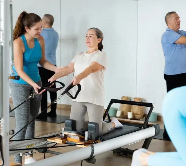 Frauen und Männer trainieren im **Reha-Sport** auf Pilatesgeräten. Eine Trainerin hilft einer älteren Frau, während ein Mann mit einem Ball übt.