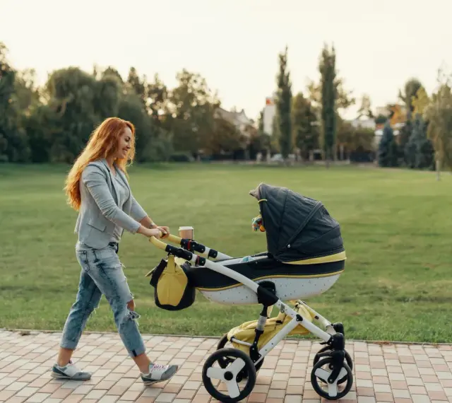Ruda kobieta z uśmiechem pcha wózek podczas pierwszego spaceru z noworodkiem w parku.