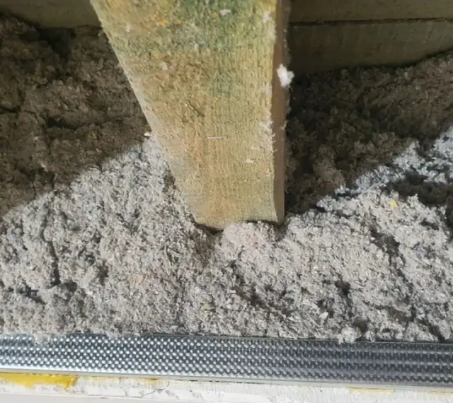 Luźna wełna celulozowa, widoczne nierówności i potencjalne wady izolacji pod drewnianą belką stropową.