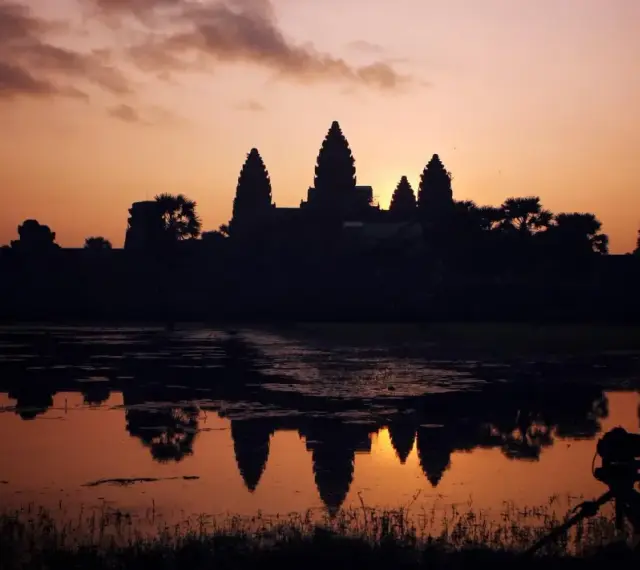 Zachód słońca nad Angkor Wat, idealne tło dla niezapomnianych kambodżańskich wakacji. Widok odbija się w wodzie, a na pierwszym planie statyw z aparatem.