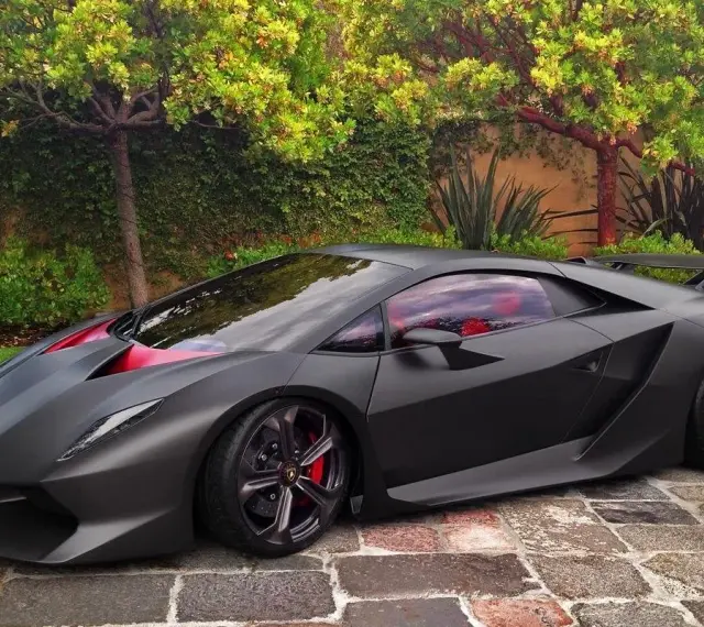 Matowy czarny Lamborghini Sesto Elemento z czerwonymi akcentami i wnętrzem, zaparkowany na brukowanej ścieżce wśród zieleni.