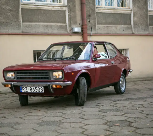 Czerwony fiat 128, klasyk z lat 70., zaparkowany na brukowanym dziedzińcu przed budynkiem.
