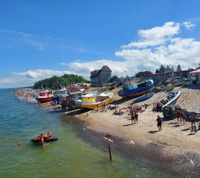 Słoneczny dzień nad morzem, plaża pełna ludzi, kolorowe łodzie na piasku. W tle widać budynki, a na wodzie pływa skuter. Idealne miejsce na wakacje, jakby ktoś przyjechał tu autobusem nad morze.