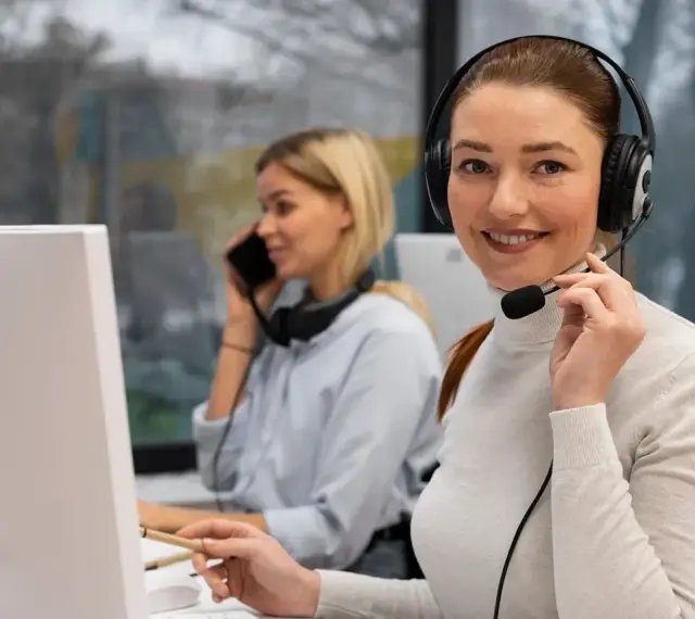 Uśmiechnięta key account manager w słuchawkach z mikrofonem pracuje przy komputerze. W tle koleżanka rozmawia przez telefon.