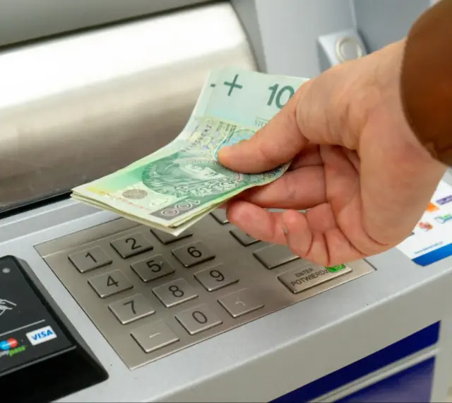 Dłoń wkłada banknoty 100 zł do bankomatu. Zastanawiasz się, ile można wpłacić na konto?