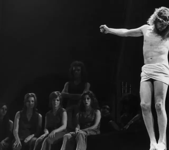 Scena z musicalu Jesus Christ Superstar: Jezus na krzyżu, otoczony przez postaci, w tym kobiety siedzące wpatrzone w niego.