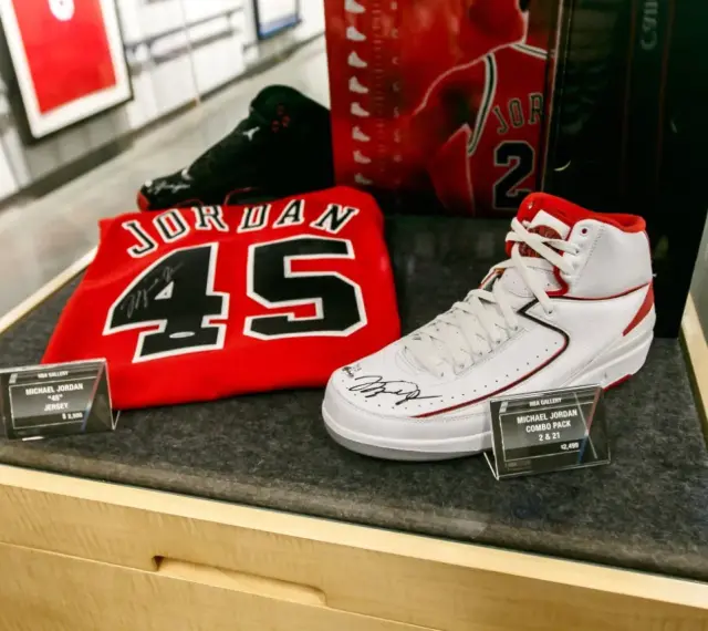Białe buty sportowe z czerwonymi akcentami, podpisane przez Michaela Jordana, obok koszulki z numerem 45. To pamiątki, które pokazują, jaki michael jordan rozmiar buta nosił.