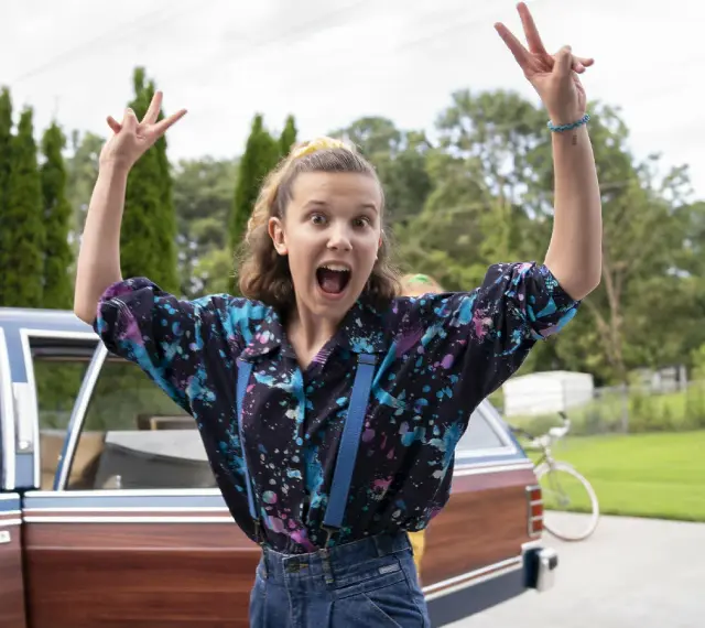 Millie Bobby Brown w kolorowej koszuli i szelkach, radośnie pozuje przy samochodzie. Jej filmy, seriale i programy to gwarancja emocji!
