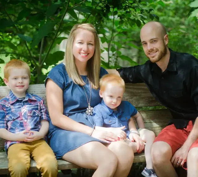 Rodzina na ławce w parku. Dwóch rudowłosych chłopców, mama i tata uśmiechają się. Młodszy syn, jakby w uporczywe naśladowanie innych dzieci, siedzi na kolanach mamy.
