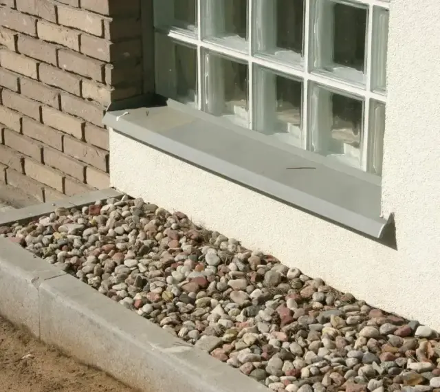 Kamienna opaska żwirowa wokół domu, otaczająca betonową rabatę z kolorowych kamieni, stanowi estetyczne i praktyczne wykończenie.