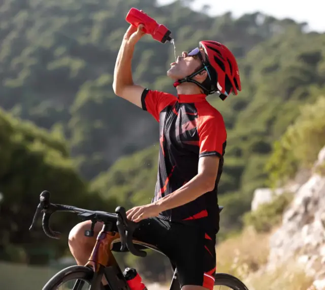 Kolarz w kasku i stroju sportowym pije wodę z bidonu, regenerując siły po wysiłku. Woda z izoleucyną pomoże mu szybciej odzyskać energię.