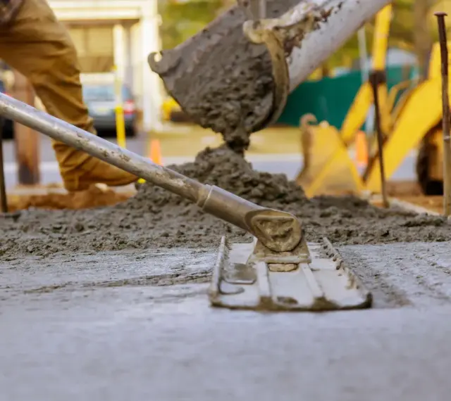 Pracownik rozprowadza świeży beton c12/15 za pomocą łaty. W tle widać koparkę i plac budowy.