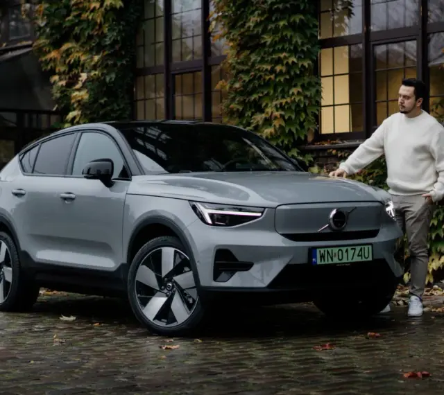 Szary Volvo C40 z czarnym dachem stoi na mokrej, brukowanej nawierzchni. Mężczyzna w białym swetrze opiera się o maskę.