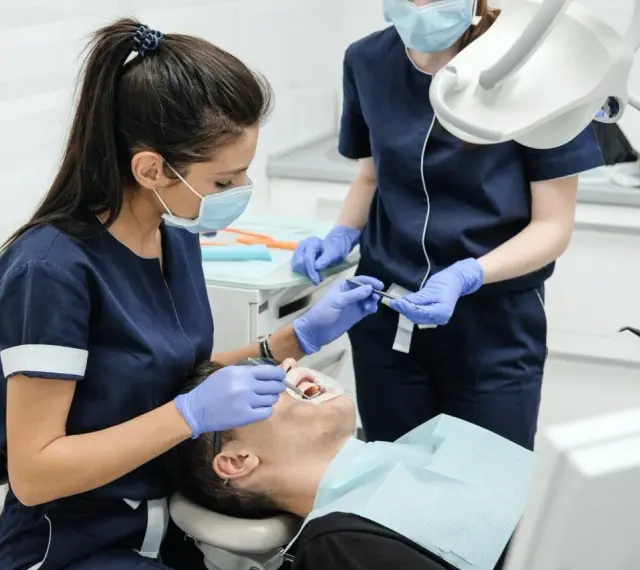Dwie dentystki w maseczkach i rękawiczkach wykonują zabieg stomatologiczny u pacjenta.
