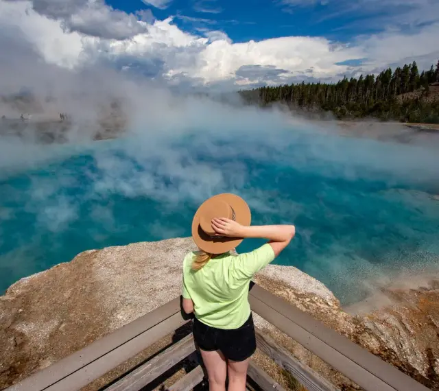 Kobieta w kapeluszu podziwia turkusowe gorące źródło w Yellowstone. Gdzie jest Yellowstone? Tu, wśród parujących wód i lasów.