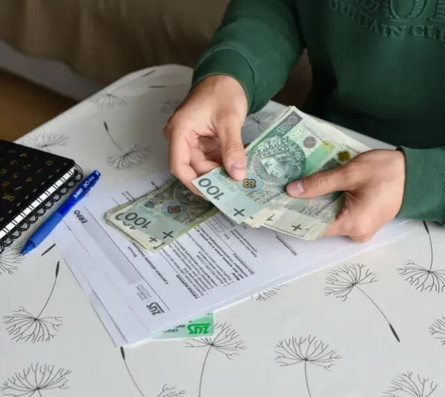 Ręce liczą banknoty 100 zł nad dokumentami ZUS. Obok leży kalkulator i długopis. To może być pomocne przy ustalaniu, ile wynosi świadczenie przedemerytalne na rękę.