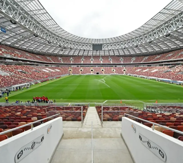 Pusta trybuna moskiewskiego stadionu z rzędem czerwonych i brązowych siedzeń. Na boisku pracują zraszacze.