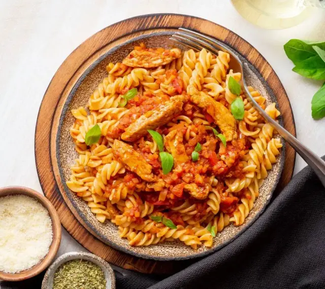 Pyszny makaron z kurczakiem w sosie pomidorowym, udekorowany bazylią. Obok miseczki z parmezanem i ziołami.
