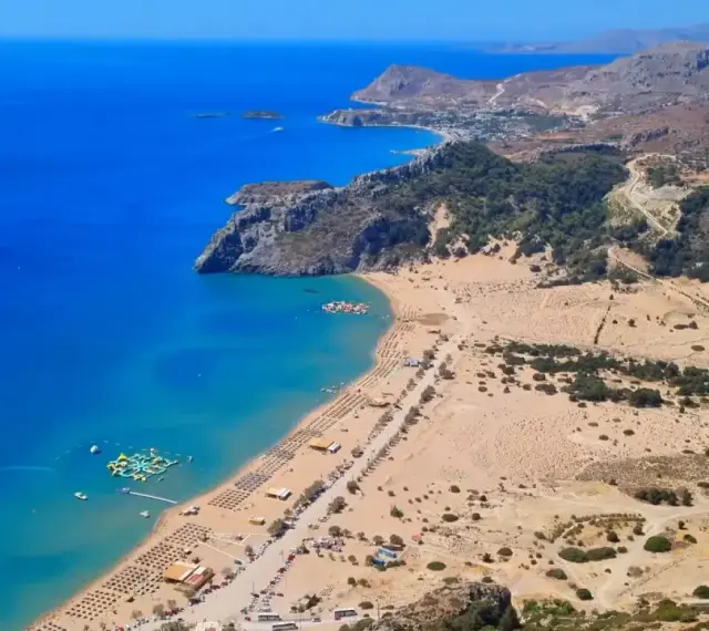 Malownicze **Rodos plaże** z turkusową wodą, piaszczystym brzegiem i zielonymi wzgórzami w tle. Widać wodny plac zabaw.