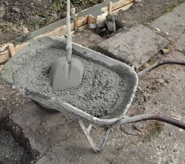 Taczka z betonem i łopatą czeka na użycie przy budowie fundamentów. W tle widać szalunki i przygotowane zbrojenie.