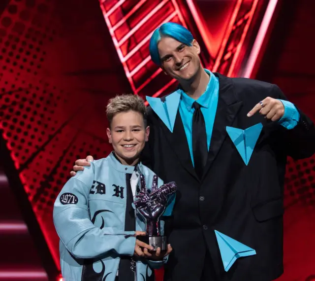 Chłopiec z błękitną kurtką i trofeum w kształcie dłoni, obok mężczyzny z niebieskimi włosami. To on wygrał The Voice Kids!