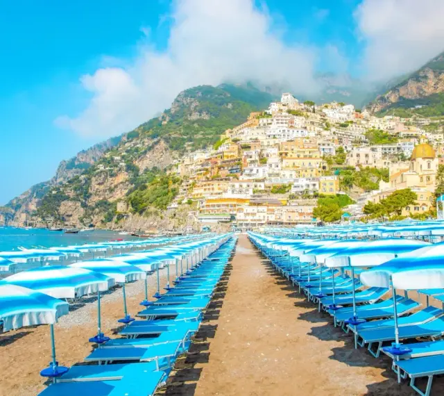 Rzędy leżaków i parasoli na plaży w Positano, Włochy. W tle widać malownicze miasteczko na zboczu góry i błękitne morze.