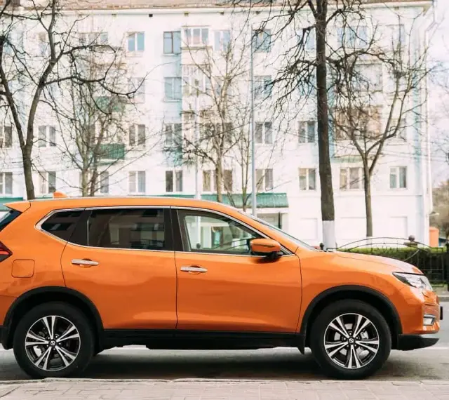 Pomarańczowy SUV do 50 tys. zaparkowany na tle białego budynku i drzew.