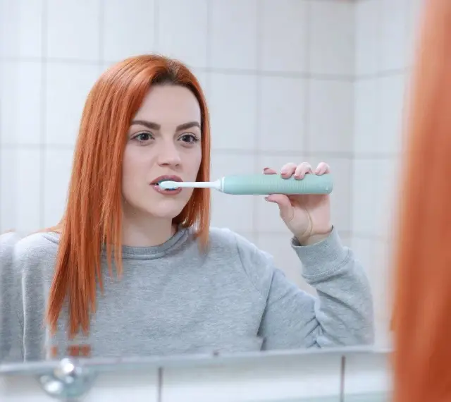 Rudowłosa kobieta myje zęby szczoteczką soniczną. Czy szczoteczka soniczna jest dobra? Ta kobieta wydaje się zadowolona.