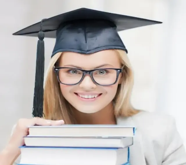 Uśmiechnięta studentka w birecie i okularach trzyma stos książek, zastanawiając się, ile trwają studia podyplomowe.