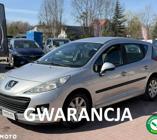 Srebrny Peugeot 207 1.4 benzyna na placu samochodowym, z napisem "GWARANCJA".