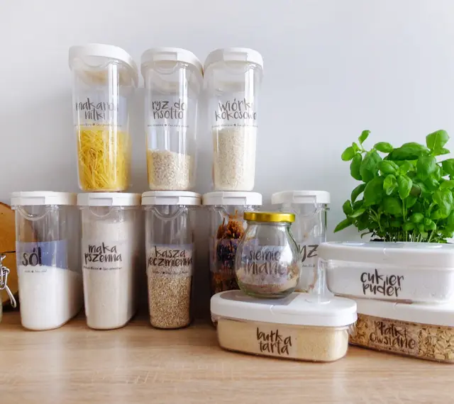 Pojemniki na żywność z etykietami pokazują, jak przechowywać pokrywki i produkty sypkie. Widać makaron, ryż, sól, mąkę, kaszę, cukier puder i płatki owsiane.