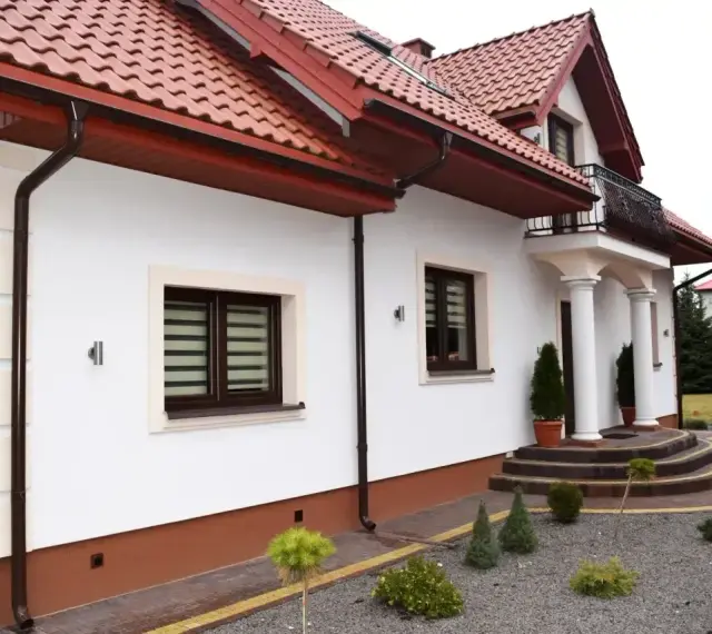 Biała elewacja domu z brązowymi okiennicami i czerwonym dachem. Przed wejściem schody i zadbany ogród.