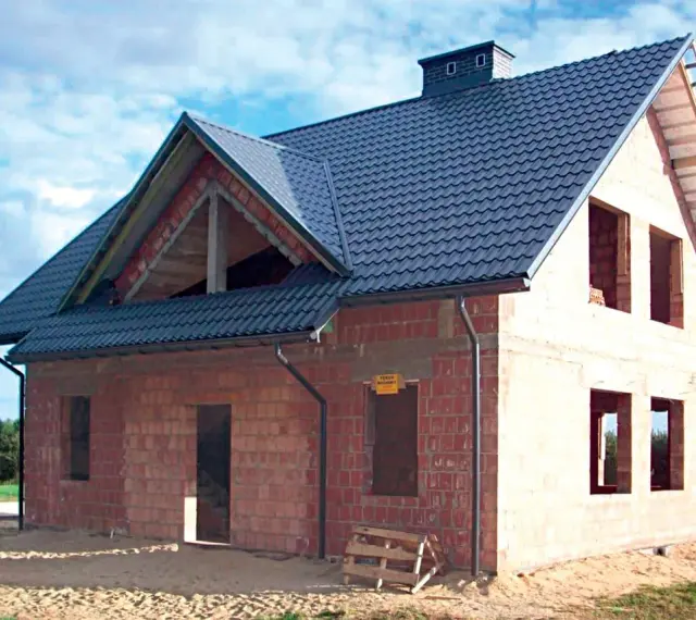 Budowa domu z cegły, widoczny dach betonowy skośny ocieplenie. Widać otwory okienne i drzwiowe, komin.