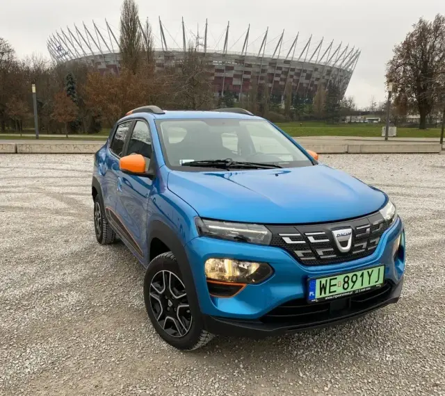 Niebieski Dacia Spring z pomarańczowymi akcentami na tle Stadionu Narodowego. Sprawdź atrakcyjną dacia spring cena!