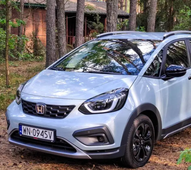 Jasny Honda Jazz Crosstar zaparkowany w lesie, obok drewnianego domku.