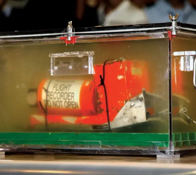 Pomarańczowa skrzynka "czarnej skrzynki" z napisem "FLIGHT RECORDER DO NOT OPEN" w przezroczystej obudowie, dowód w sprawie wypadku lotniczego.