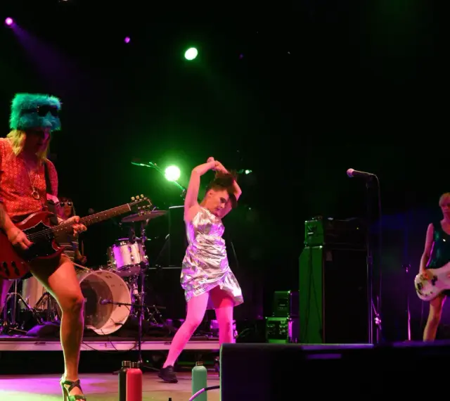 Sur scène, le groupe Bikini Kill enflamme le public. Une guitariste porte un chapeau vert fluo, une autre joue de la basse, et une troisième danse avec énergie.