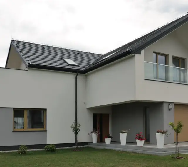 Nowoczesna elewacja budynku z szarymi i białymi tynkami, dużym oknem, balkonem i drewnianą bramą garażową.