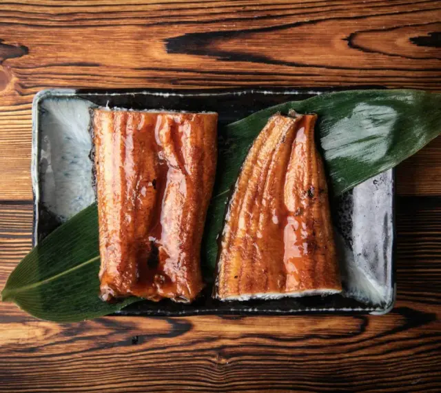 Dwa kawałki błyszczącego, glazurowanego sushi z węgorzem na liściu, podane na czarnej tacce.