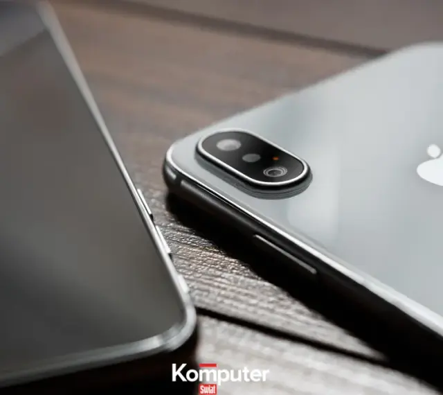 Dwa telefony leżą obok siebie na drewnianym blacie. Jeden z nich, srebrny iPhone, ma widoczny moduł aparatu. Drugi, z ciemnym ekranem, jest ustawiony bokiem. Idealne do rozmowy przez **Facetime**.