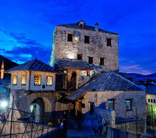 Kamienne budynki i most w Mostarze o zmierzchu, z widocznym minaretem w tle.