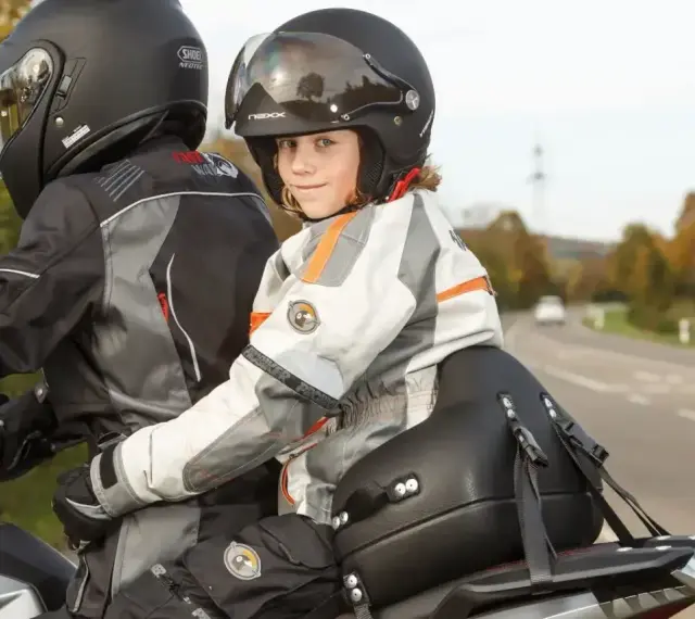 Uśmiechnięte dziecko na motocyklu, siedzące przed dorosłym, w kasku i kurtce motocyklowej.