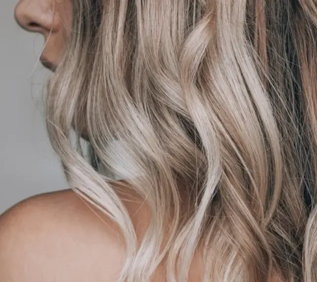 Kobieta z falowanymi, blond włosami ozdobionymi nowoczesnymi pasemkami, ukazana od ramienia.