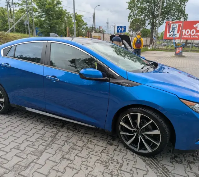 Błękitny Chevrolet Volt z czarnym dachem, zaparkowany na mokrej kostce brukowej. W tle widać stację KFC.
