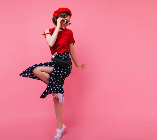 Dziewczyna w czerwonym beretce i sukience w grochy, w stylu pin up, skacze na różowym tle.