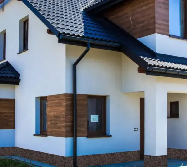 Nowoczesny dom z białym tynkiem i drewnianymi akcentami. Z czego zrobić cokół wokół domu? Widać tu kamyczki, które mogą być inspiracją.