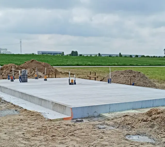 Gotowa płyta fundamentowa na budowie, otoczona piaskiem i zielenią. W tle widać pola i budynki przemysłowe.
