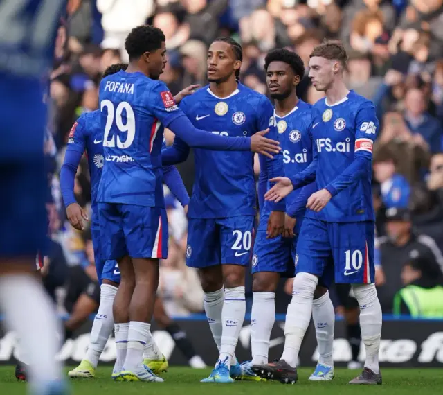 Piłkarze Chelsea w niebieskich strojach rozmawiają na boisku. Widać numery 29, 20 i 10.