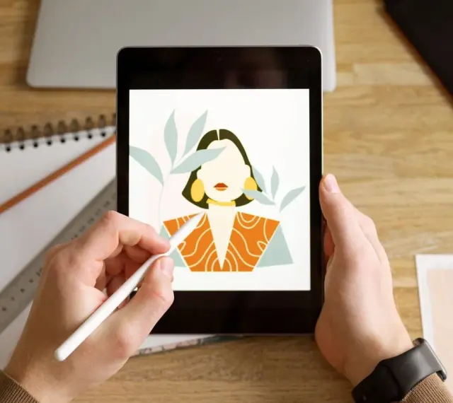 Ręce trzymają tablet z rysunkiem kobiety, tworzonym w aplikacji procreate. Obok leżą szkicownik i ołówek.